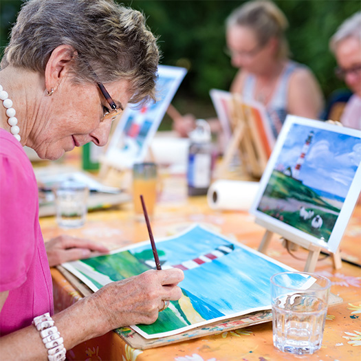 A senior woman painting at an outdoor table.