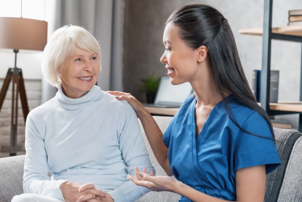 A nurse converses with a senior woman on a couch.