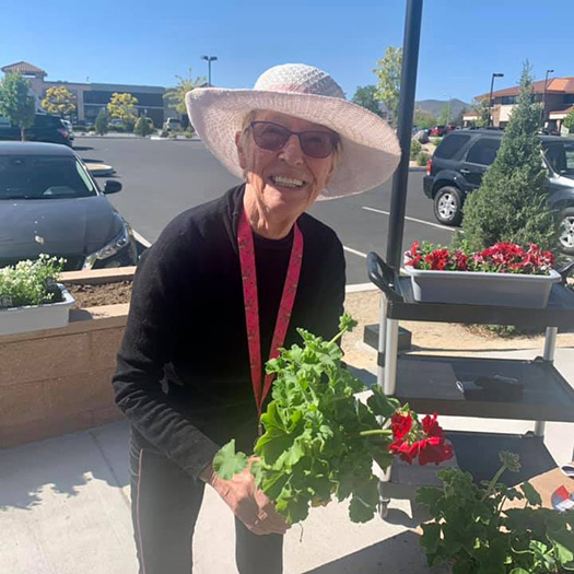 A senior woman in a hat holding a plant in front of the senior home.